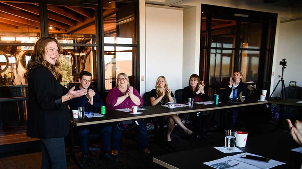 Presentation Skills Training Orange County, California 2 A woman stands and speaks to a group of people seated at a conference table, who are smiling and clapping in a modern meeting room—a dynamic scene from Presentation Skills and Public Speaking Training.